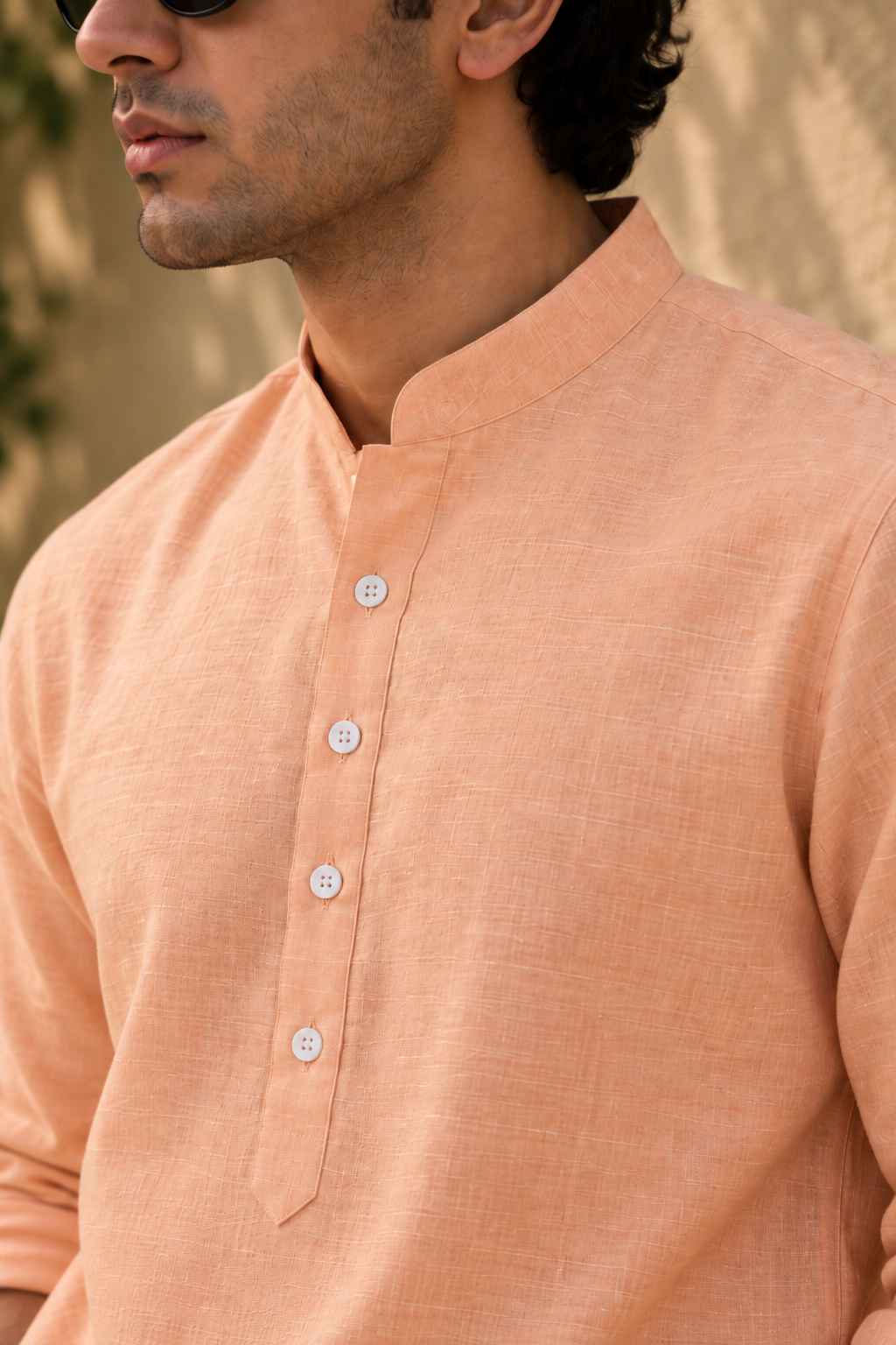 A close-up of a man wearing a peach-colored linen kurta with a mandarin collar and white buttons, set against a warm, blurred background.