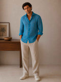 Man wearing a blue shirt and beige pants standing in a room with a wooden table and framed picture.