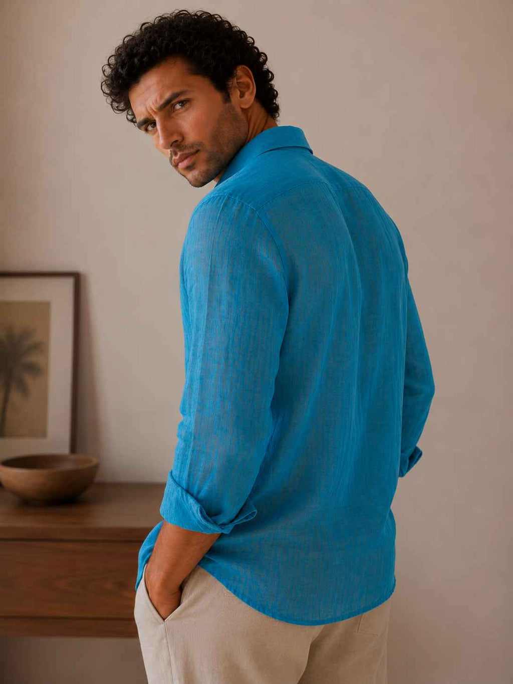 Man wearing a blue shirt standing in a room with a wooden shelf and framed picture.