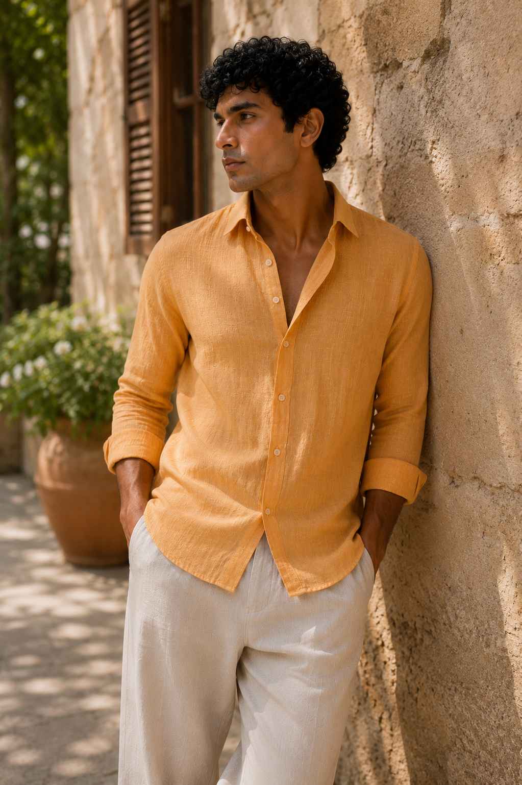 A man wearing a mustard yellow linen button-up shirt and off-white trousers, leaning against a rustic stone wall outdoors.