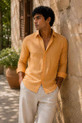 A man wearing a mustard yellow linen button-up shirt and off-white trousers, leaning against a rustic stone wall outdoors.