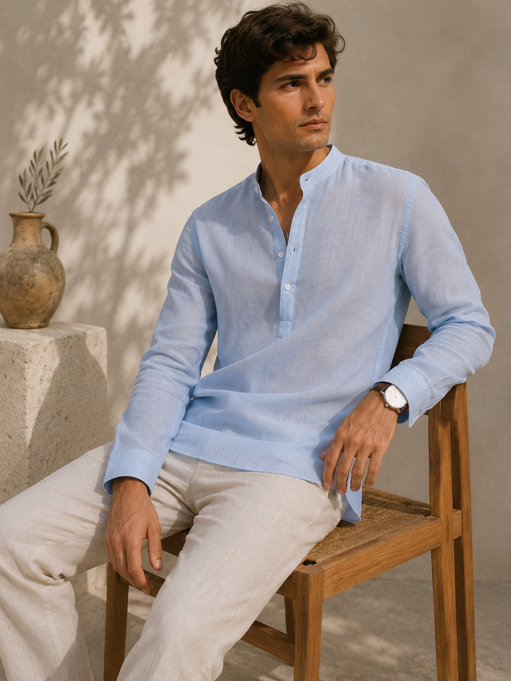 Man sitting on a wooden chair wearing a light blue shirt and beige pants against a neutral background.