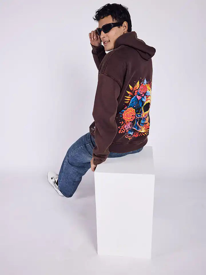 A person wearing a brown hoodie with a flowery skull print on the back, sitting on a white cube.